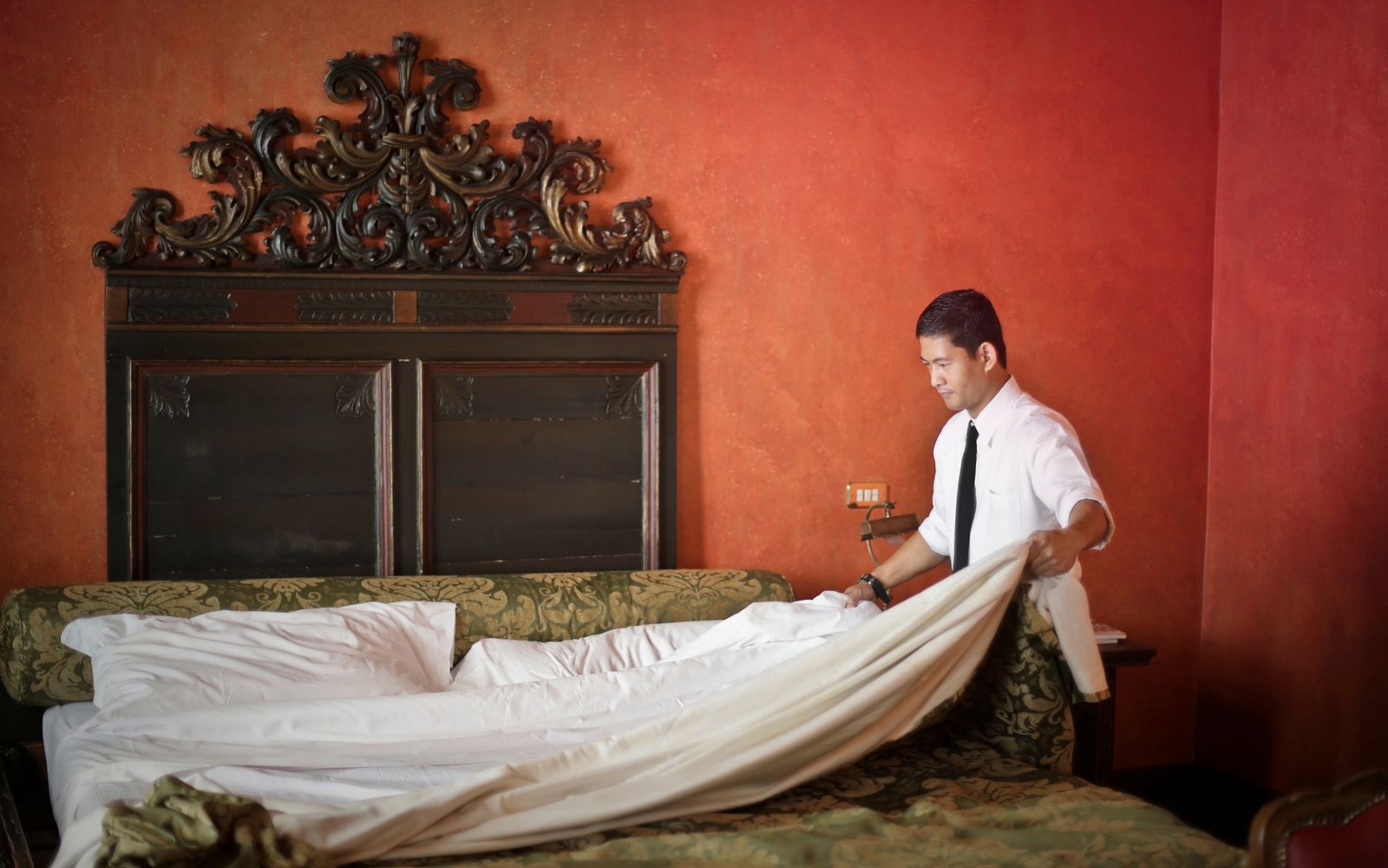 Hotel staffer in white shirt makes a bed with an ornately carved wooden headboard