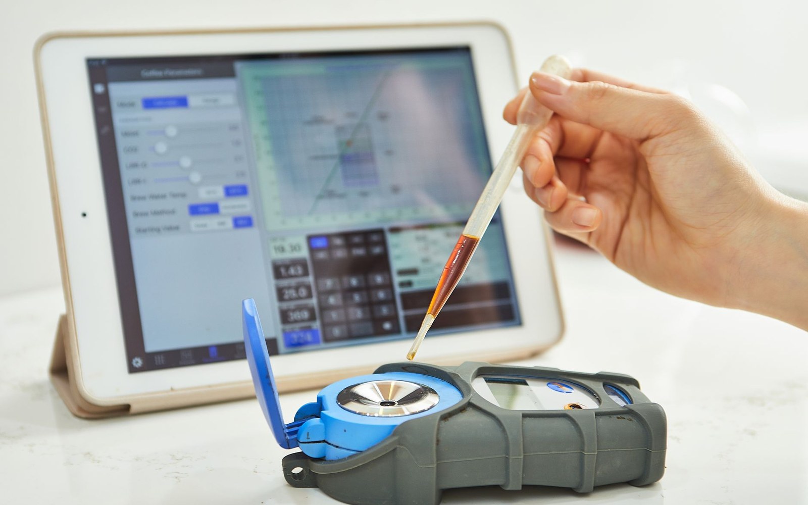 A hand drops liquid from a pipette into a refractometer beside a tablet showing coffee data