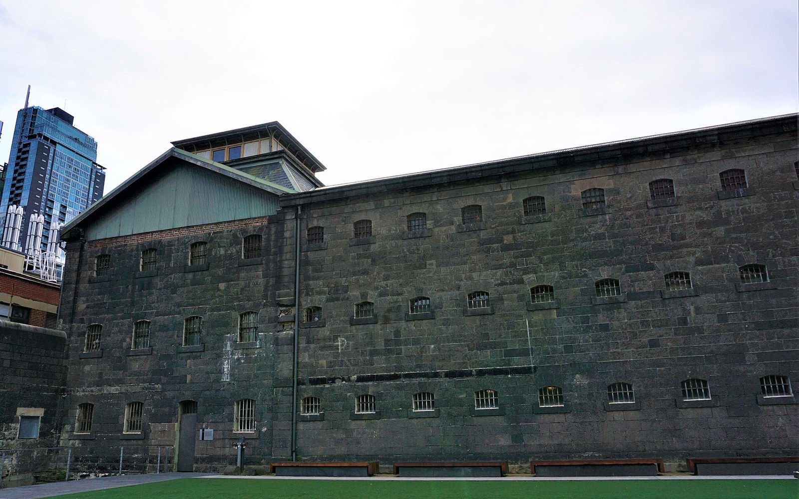 Old bluestone prison building with barred windows stands beside a modern skyscraper