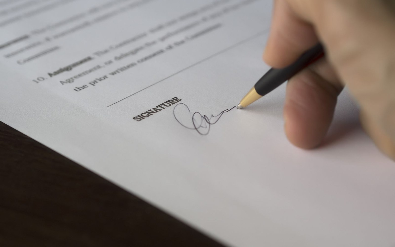 A hand signs a document with a pen above the printed word "signature"