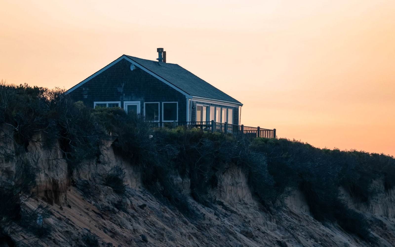 Shingled cottage with a deck perches atop an eroding coastal bluff at dusk
