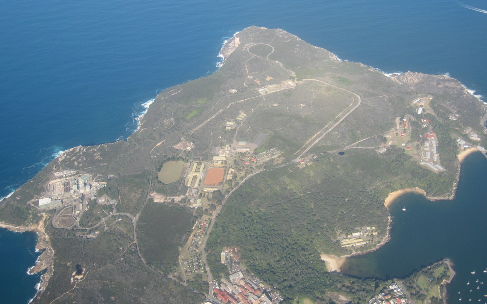 Aerial view of a coastal headland with scattered buildings, roads and sports courts amid bushland