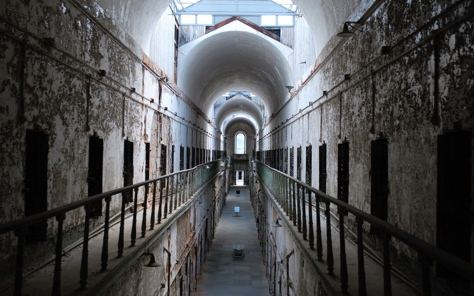 A long cellblock corridor with peeling paint and tiered iron balconies under an arched skylight