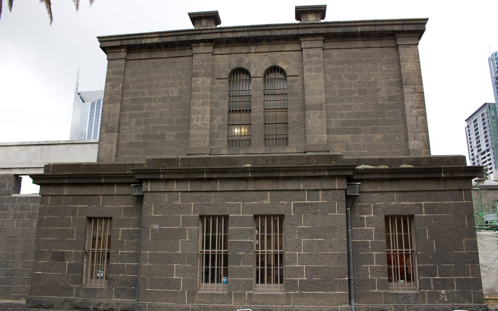 A corridor inside Old Melbourne Gaol, cell doors open on both sides, available light from a high window, an adult visitor visible at the far end