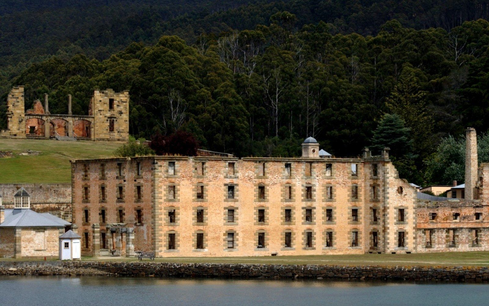 The Port Arthur penitentiary ruin at dusk, stone walls open to the sky, an adult tour guide in the foreground holding a lantern